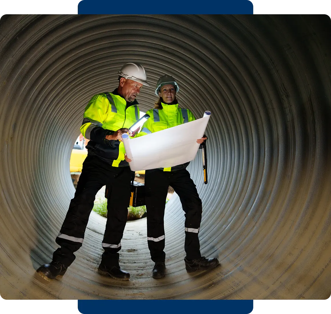 Two workers in safety gear reviewing plans inside a large pipe tunnel.