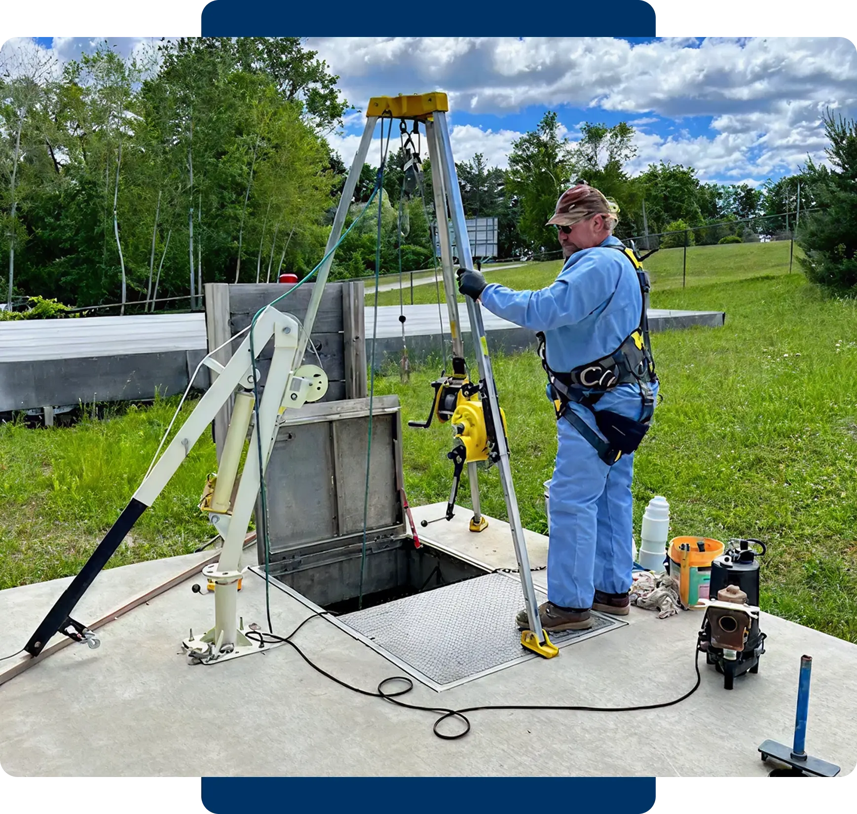 A man operates a tripod hoist lifting a large object outdoors.