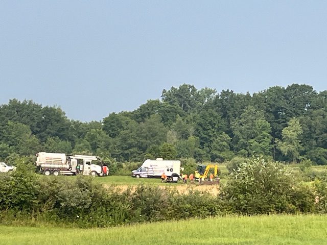 Construction vehicles and trucks near a forested area under a clear sky.